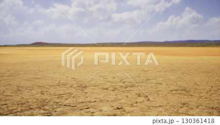 A vast area of cracked yellowish earth stretches across the landscape, with sparse greenery visible in the distance under a bright blue sky adorned with fluffy clouds. A vast area of cracked yellowish earth stretches across the landscape, with sparse greenery visible in the distance under a bright blue sky adorned with fluffy clouds. 130361418