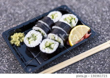 A tray holds several fresh cucumber sushi rolls, paired with a dollop of wasabi and a slice of lemon. The setting features a textured surface, emphasizing the vibrant colors of the dish A tray holds several fresh cucumber sushi rolls, paired with a dollop of wasabi and a slice of lemon. The setting features a textured surface, emphasizing the vibrant colors of the dish 130361723