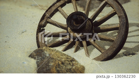 A weathered wooden wagon wheel lies partially buried in sand beside a large stone. The seaside landscape features a gentle slope of sand with hints of a coastal environment. 130362230