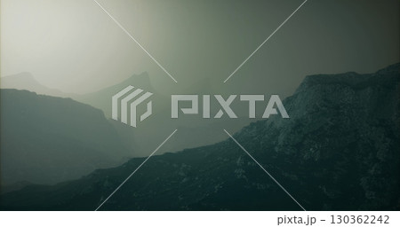 Fog envelops the rugged mountain range during early morning hours, revealing silhouettes of peaks and rocky formations in a serene, yet ethereal atmosphere. 130362242