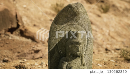 A detailed stone sculpture showcases a figure, partially hidden by a weathered surface, set against a sandy background under bright sunlight, hinting at ancient artistry and culture. A detailed stone sculpture showcases a figure, partially hidden by a weathered surface, set against a sandy background under bright sunlight, hinting at ancient artistry and culture. 130362250