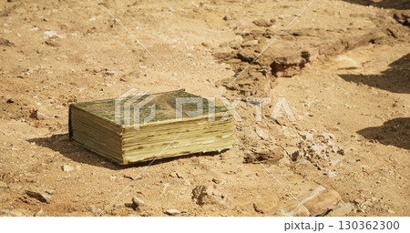 An aged book rests on dry, cracked earth surrounded by rocky terrain. Sunlight illuminates its weathered pages, hinting at stories long forgotten in this desolate location. 130362300