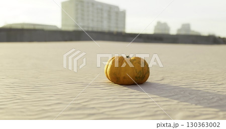 A vibrant orange pumpkin sits alone on a sunlit sandy landscape. Its textured surface contrasts with smooth grains, creating a striking focal point during the quiet dusk. 130363002
