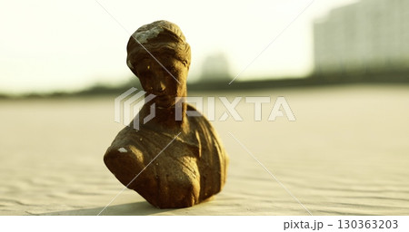 A weathered bronze bust of a woman stands prominently amidst the golden sand, illuminated by soft rays of the setting sun. Gentle waves lap at the shore nearby. 130363203