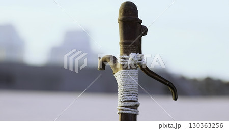 An old water pump, weathered and worn, stands amidst a tranquil landscape. Twine wraps around its body, hinting at years of service. This relic embodies history and simplicity, evoking nostalgia. 130363256