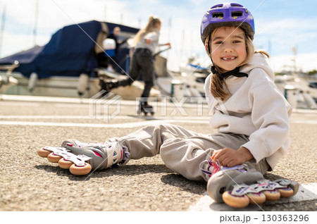 Happy child in protective gear on rollerblades sitting by boats at harbor Happy child in protective gear on rollerblades sitting by boats at harbor 130363296