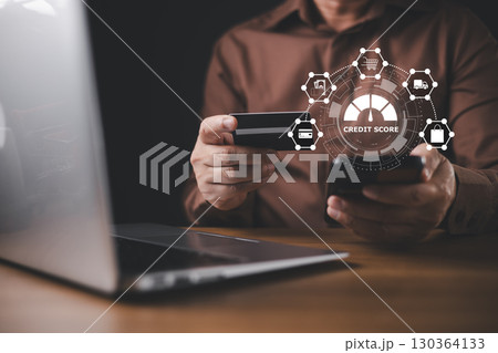 With a hologram displaying a credit score report, a businessman uses a credit card and laptop to conduct financial analysis and manage online payments through advanced technology. 130364133