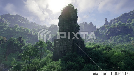 A towering rock formation stands prominently amidst a dense forest, illuminated by sunlight filtering through the clouds. The mountainous landscape showcases vibrant foliage under a clear sky. 130364372