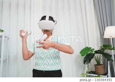 Asian girl wearing VR headset, playing an immersive video game with controllers in a modern room. 130364391
