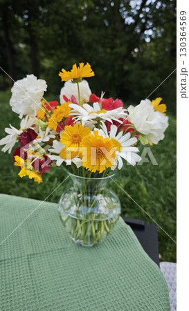 Mixed bouquet of summer flowers in a clear glass vase placed on a green tablecloth outdoors 130364569
