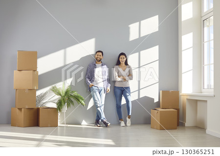 Happy family couple in casual clothes are standing together near wall in rays of sunlight among cardboard boxes. Married smiling couple buys or rents new house. Real estate, relocation, own housing. 130365251