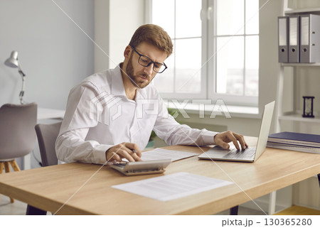 Focused businessman working on accounts using a calculator and laptop Focused businessman working on accounts using a calculator and laptop 130365280