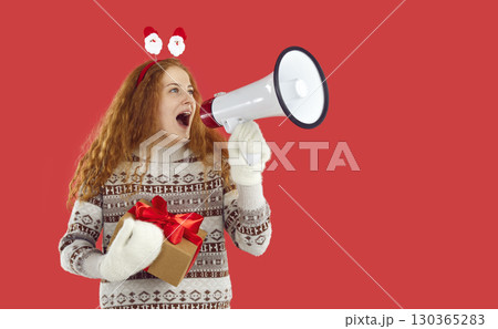 Happy redhead girl holding Christmas gift present in hands shouting loudspeaker on red background. Happy redhead girl holding Christmas gift present in hands shouting loudspeaker on red background. 130365283