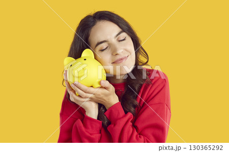 Smiling young woman gently leans her cheek to piggy bank, isolated on orange background. Smiling young woman gently leans her cheek to piggy bank, isolated on orange background. 130365292