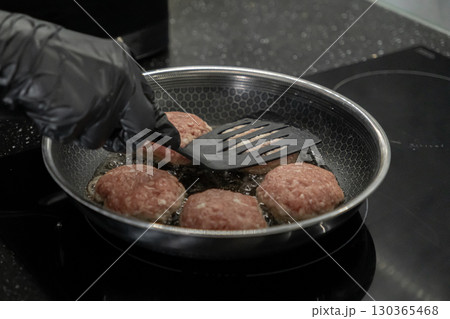 Person in black glove frying raw cutlets with a spatula in a hot pan on an induction stove for home cooking, healthy meal preparation 130365468