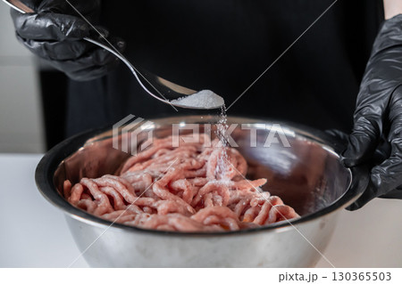 Professional chef's gloved hand carefully sprinkles white salt from a spoon onto raw minced meat in a metal bowl, preparing fresh homemade cutlets 130365503