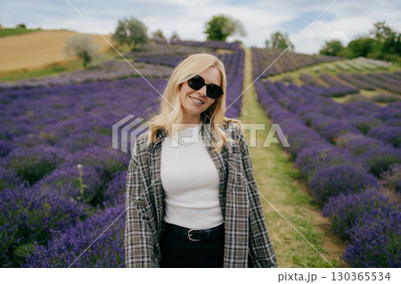 Stylish young woman wearing sunglasses is smiling while standing in vibrant lavender field, enjoying beautiful scenery and fragrance of blooming flowers. Fashionable woman stands amid lavender blooms 130365534