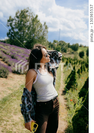 Woman relaxing in lavender field, wearing sunglasses, casual attire, capturing scenic landscape with smartphone while enjoying warm sunlight and fragrant surroundings. Woman posing in lavender field Woman relaxing in lavender field, wearing sunglasses, casual attire, capturing scenic landscape with smartphone while enjoying warm sunlight and fragrant surroundings. Woman posing in lavender field 130365537