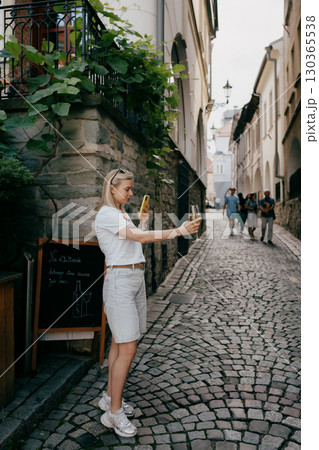 Young blonde woman taking picture of glass of white wine with her smartphone in narrow cobbled street of old town. Blonde woman photographing white wine glass near historic cobblestone alley 130365538