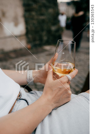Woman sitting outdoors in casual attire, savoring glass of chilled white wine while enjoying peaceful moment of relaxation amidst vibrant urban backdrop. Woman relaxing outdoors, holding white wine Woman sitting outdoors in casual attire, savoring glass of chilled white wine while enjoying peaceful moment of relaxation amidst vibrant urban backdrop. Woman relaxing outdoors, holding white wine 130365543