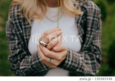 Close up of woman gently holding single flower stem, showcasing her ring and plaid shirt against blurred natural background, evoking themes of nature, femininity, and simple beauty. Thin stem in hands Close up of woman gently holding single flower stem, showcasing her ring and plaid shirt against blurred natural background, evoking themes of nature, femininity, and simple beauty. Thin stem in hands 130365548
