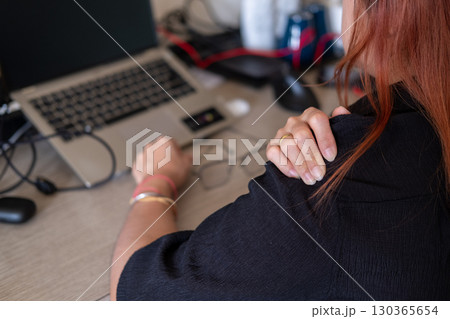 Woman massaging sore shoulders at desk with laptop and office tools highlighting work fatigue 130365654