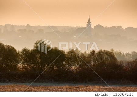 rural landscape on an autumn morning. row of trees along the empty fields. village with church on the hill behind the forest in the hazy distance rural landscape on an autumn morning. row of trees along the empty fields. village with church on the hill behind the forest in the hazy distance 130367219