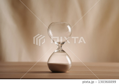 A simple hourglass on a wooden table with a beige backdrop, symbolizing time flow and minimalistic living. Represents timeless themes in a creative, uncluttered presentation for stylish concepts. 130368899