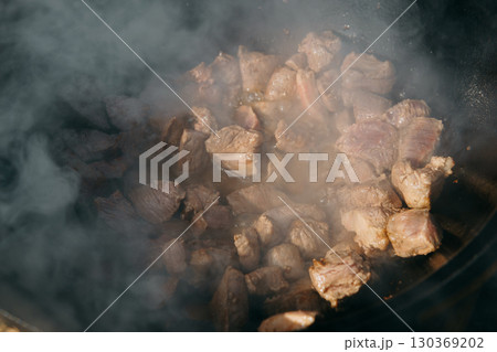Pieces of meat frying in oil in a large cast iron cauldron with steam. Cooking outdoor in a big pot for holiday. Pieces of meat frying in oil in a large cast iron cauldron with steam. Cooking outdoor in a big pot for holiday. 130369202