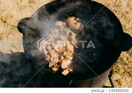Pieces of meat frying in oil in a large cast iron cauldron with steam. Cooking outdoor in a big pot for holiday. Pieces of meat frying in oil in a large cast iron cauldron with steam. Cooking outdoor in a big pot for holiday. 130369203