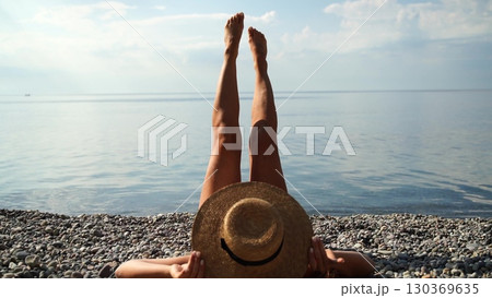 Beach Sunbathing Woman: Woman relaxes on pebble beach, legs up, sunny day by the sea, enjoying vacation. 130369635