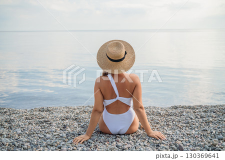 A woman wearing a white bikini and a straw hat is sitting on a beach. The beach is rocky and the sky is cloudy. 130369641