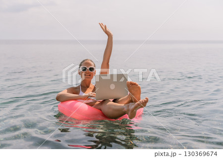 A woman is sitting on a pink inflatable raft in the ocean with a laptop on her lap. She is smiling and she is enjoying her time. A woman is sitting on a pink inflatable raft in the ocean with a laptop on her lap. She is smiling and she is enjoying her time. 130369674