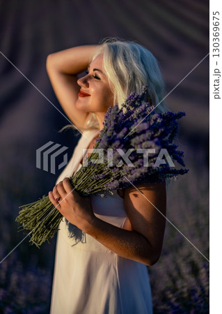 Blonde woman poses in lavender field at sunset. Happy woman in white dress holds lavender bouquet. Aromatherapy concept, lavender oil, photo session in lavender Blonde woman poses in lavender field at sunset. Happy woman in white dress holds lavender bouquet. Aromatherapy concept, lavender oil, photo session in lavender 130369675