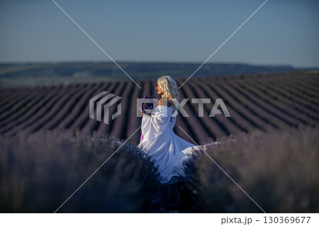 Blonde woman poses in lavender field at sunset. Happy woman in white dress holds lavender bouquet. Aromatherapy concept, lavender oil, photo session in lavender Blonde woman poses in lavender field at sunset. Happy woman in white dress holds lavender bouquet. Aromatherapy concept, lavender oil, photo session in lavender 130369677