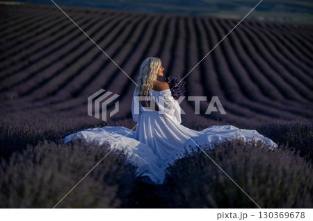 Blonde woman poses in lavender field at sunset. Happy woman in white dress holds lavender bouquet. Aromatherapy concept, lavender oil, photo session in lavender 130369678