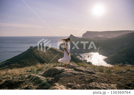woman stands on a rocky hill overlooking a body of water. She is wearing a white dress and she is in a state of joy or celebration. Concept of freedom and happiness. 130369764