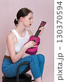 Serious young female musician playing ukulele, sitting on bar stool and looking away while plucking musical instrument. Profile view slender woman in casual clothing - white vest, blue skinny jeans 130370594
