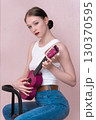 Woman musician is sitting and playing ukulele, her face expresses serenity as she looking at camera. Elegant young woman strumming stringed musical instrument in her casual, comfortable outfit 130370595