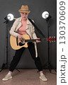Confidence young female guitarist with Spanish six string acoustic guitar standing full length on stage and looking at camera. Musician dressed in beige leather jacket, straw hat, black jeans pants 130370609