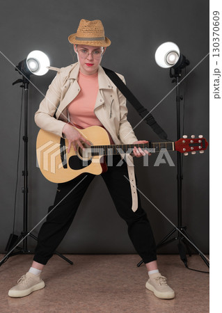 Confidence young female guitarist with Spanish six string acoustic guitar standing full length on stage and looking at camera. Musician dressed in beige leather jacket, straw hat, black jeans pants 130370609