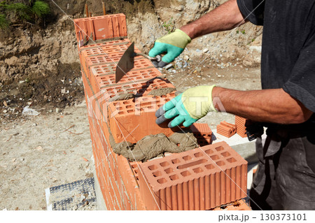 Skilled bricklayer constructs a sturdy wall using red bricks and mortar under bright sunlight in a construction site 130373101