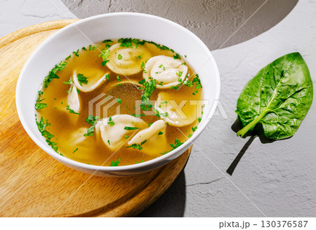Warm dumpling soup served in a white bowl with herbs and spinach on wooden board 130376587
