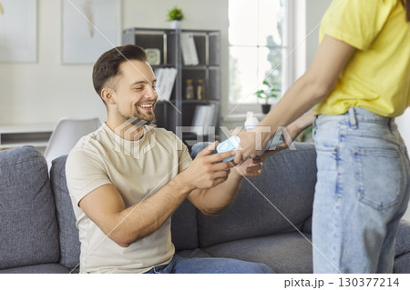 Young woman holding present gift box in hand congratulating her boyfriend sitting on sofa at home. Young woman holding present gift box in hand congratulating her boyfriend sitting on sofa at home. 130377214