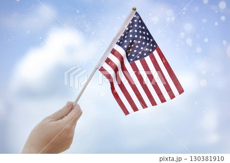 Human hand holding an American flag with a blue sky background. 4th of July concept. Close up on USA flag in a hand of a person. Independence Day or traveling in America concept. 130381910