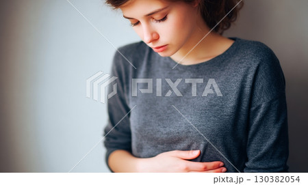 A young woman clutches her stomach, showing signs of discomfort in a softly lit room 130382054