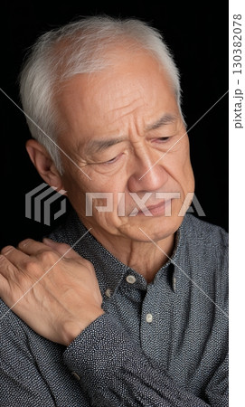 Senior man shows signs of discomfort, holding his shoulder with a thoughtful expression in a dim setting 130382078