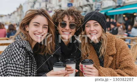 Three smiling women enjoying coffee outdoors in cozy winter attire, showcasing friendship and warmth in a lively urban setting 130382207