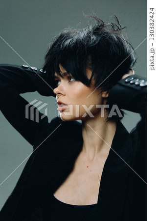 Stylish woman with short black hair in a black jacket posing against a dark background, showcasing modern fashion and confident expression. 130382424