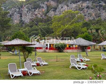 Sun loungers with straw umbrellas on a green lawn against mountain cliffs. Leisure, travel and relaxation in a natural resort environment. Sun loungers with straw umbrellas on a green lawn against mountain cliffs. Leisure, travel and relaxation in a natural resort environment. 130382477